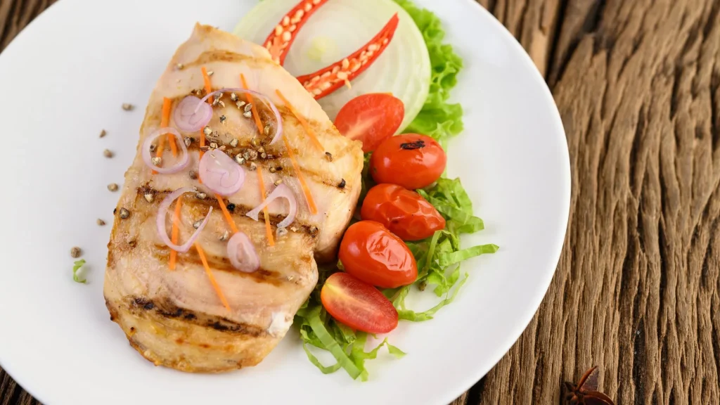 Grilled chicken on a white plate with salad, tomatoes, onion, chili, and sauce